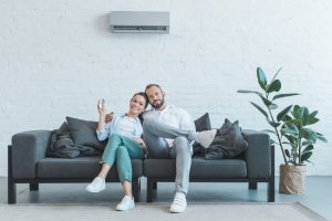One half of a couple aims a remote at an air conditioner as they both sit on a couch smiling.